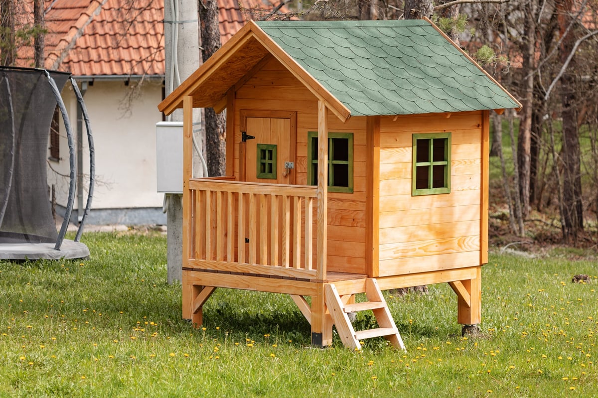 A small wooden playhouse with green roof, balcony, and stairs in a backyard